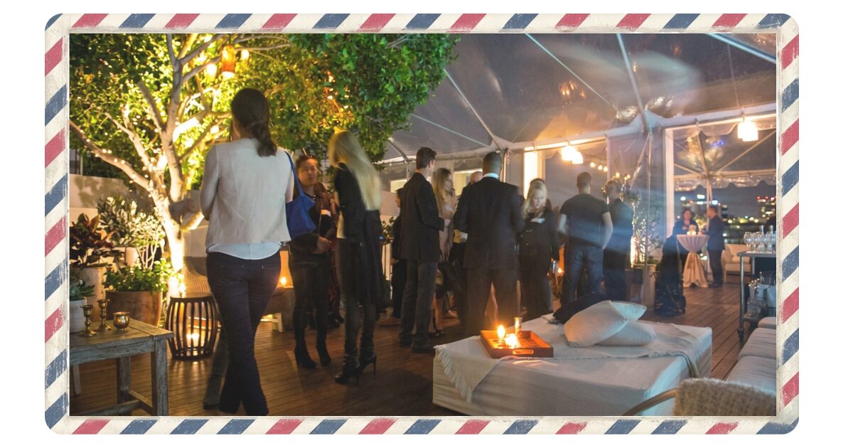 複数の人が立って談笑している集まり。英語を話す人々が集まった夜間、室内のパーティイベント会場、
様々な国籍の人々が話し合っている